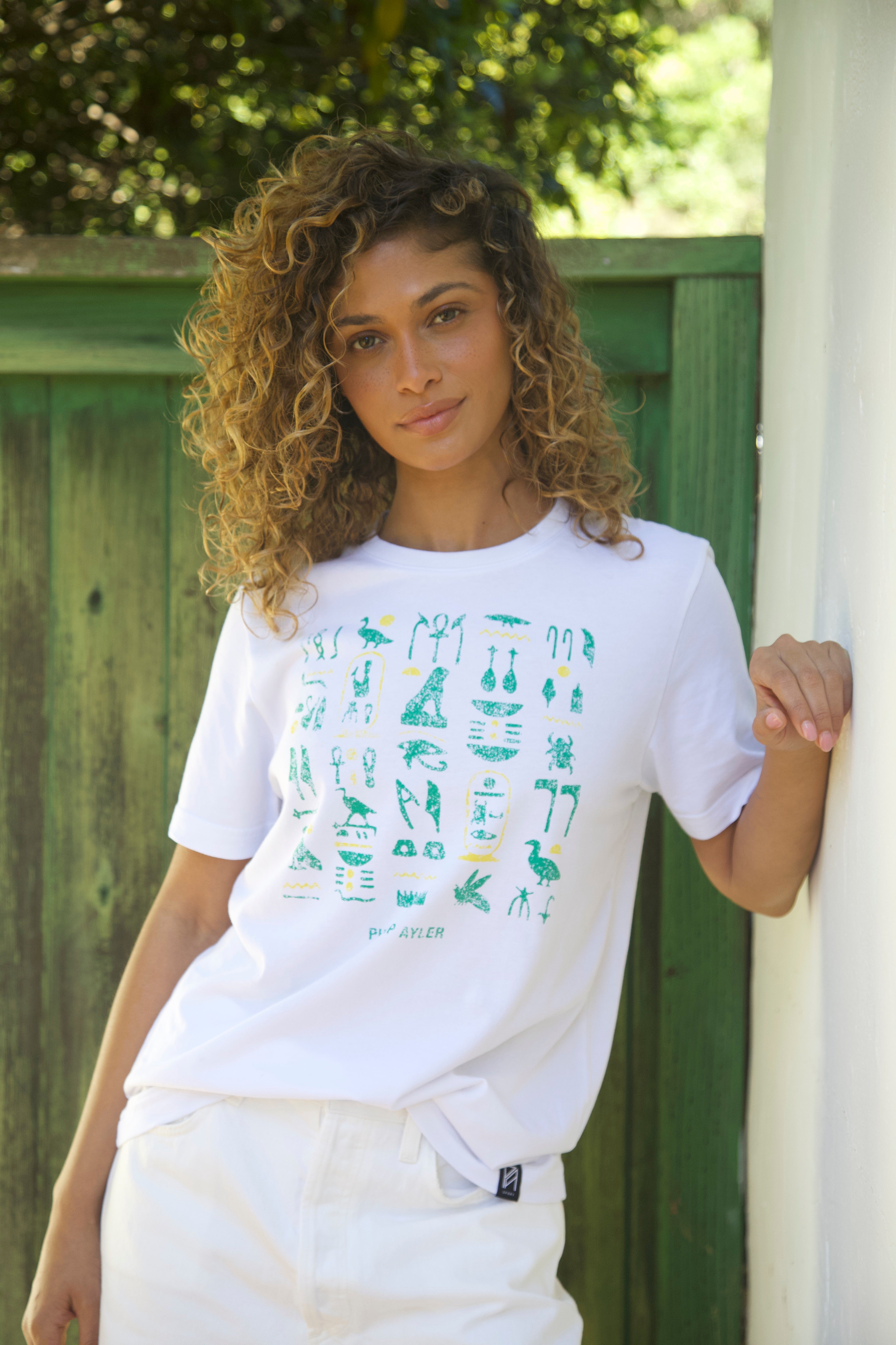 A woman with curly brown hair wearing a white t-shirt with a green hieroglyphic graphic print, posing outdoors against a green wooden fence.