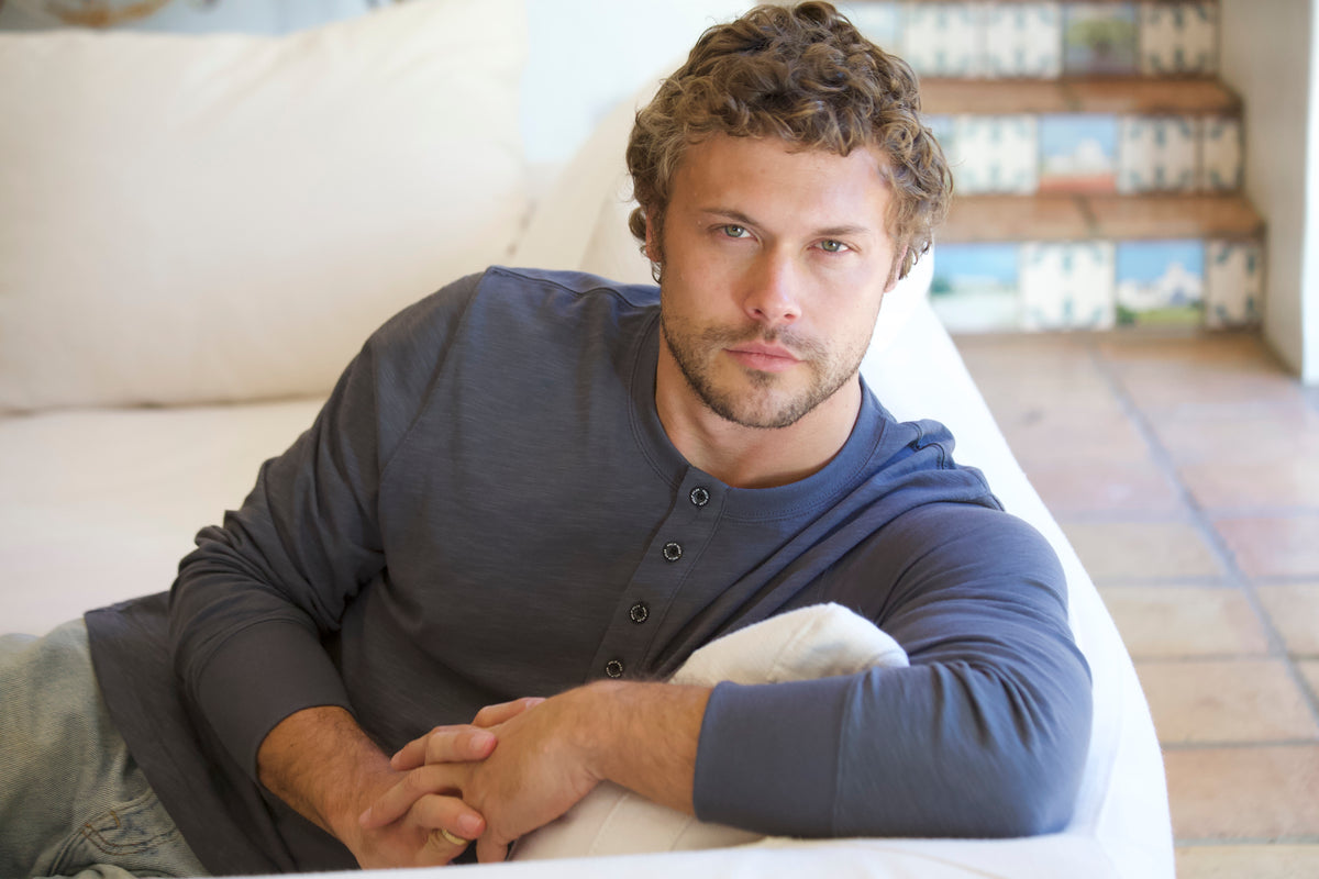 A man with curly brown hair and a beard wearing a dark gray long-sleeve henley shirt, lying on a white sofa in a brightly lit room.