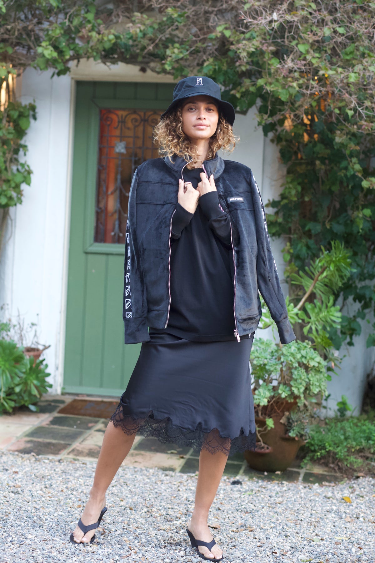 A woman stands outdoors in front of a white house with a green door, wearing a black bucket hat, black jacket, black top, black skirt, and black flip-flops.